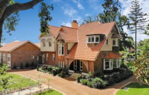 Exterior shot of a federation, arts and crafts-style heritage house. Made out of brick, terracotta tiles, painted white timber window and door frames, some bay windows, and a neat, well-maintained garden. 