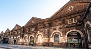 Exterior shot of the Locomotive Workshops at South Everleigh, numbers 10-15 in view. The windows are all curved and arched, with pointed, pitched roofs, and remnants of the old tracks in the ground. Inside are shops, offices, steel and metal workshops, and cafes and restaurants. 