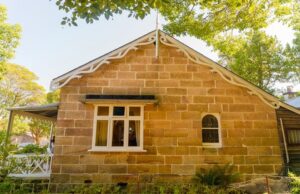 Medium-close up, side exterior shot of the Vienna Cottage in Hunter’s Hill, NSW. Sandstone cottage with small, timber framed windows, pitched roof, and front veranda.